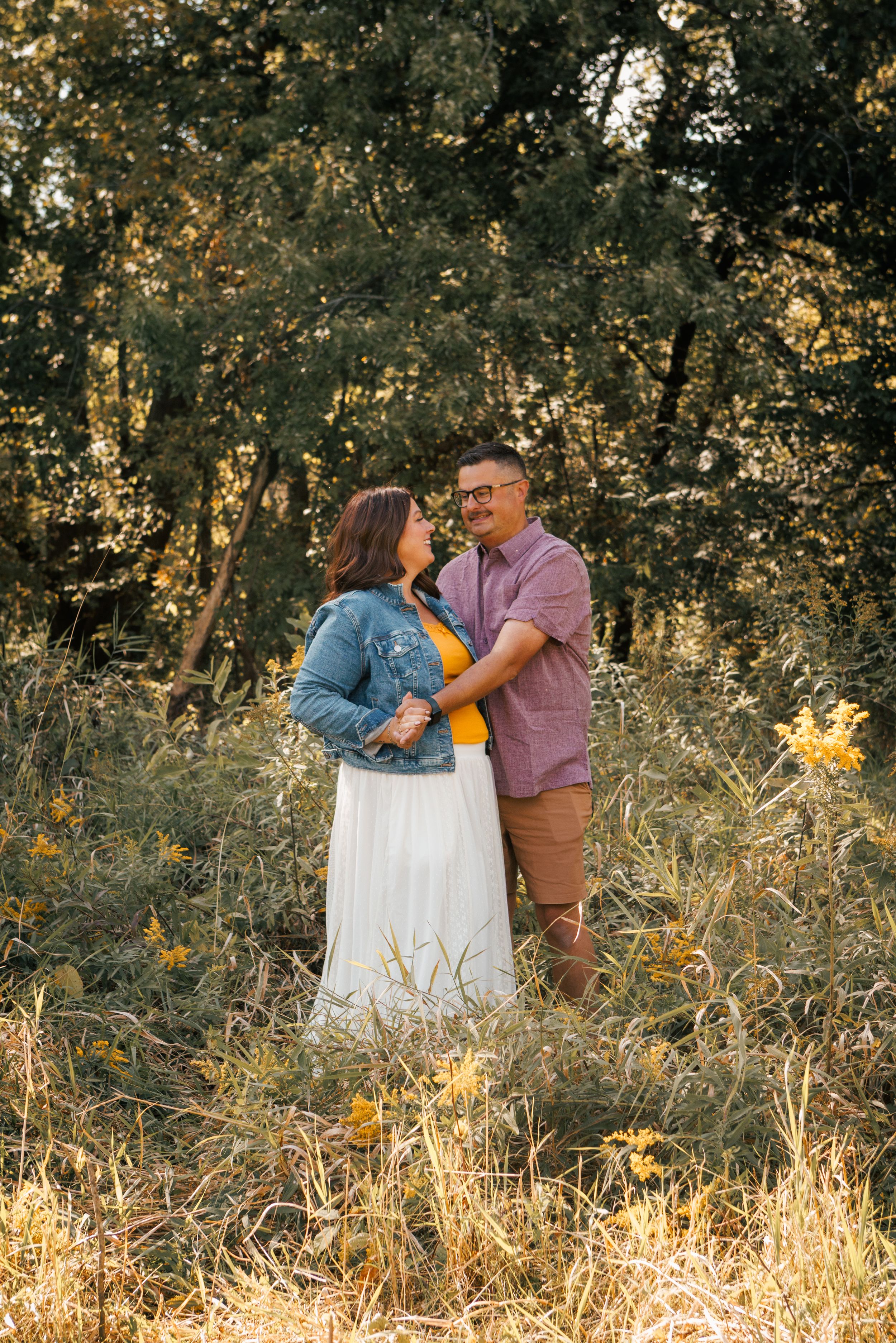 Couple embracing in golden field with lush green trees, woman in white skirt and denim jacket, man in pink shirt.