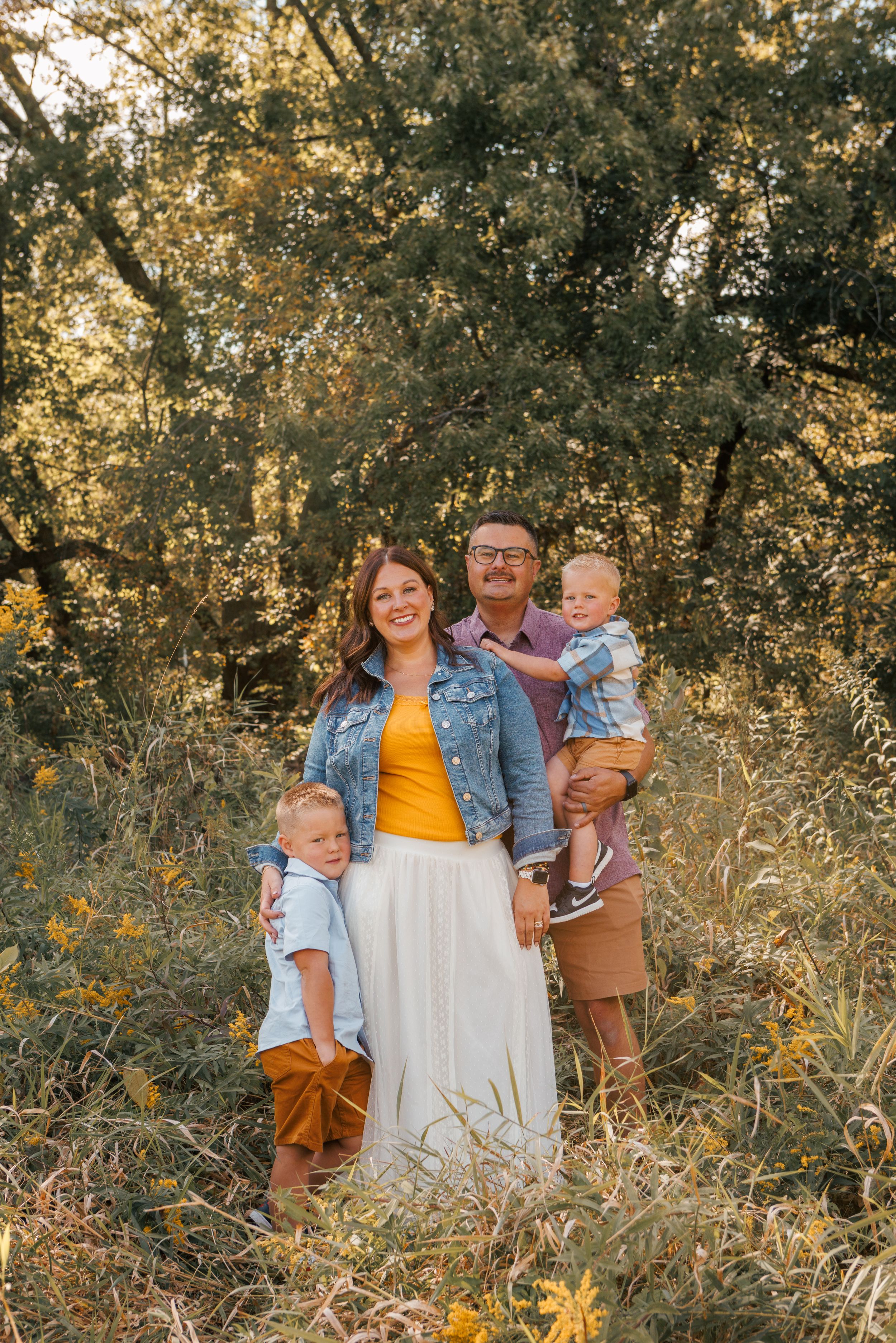 A happy family of four poses outdoors in golden light, surrounded by lush greenery and wildflowers.