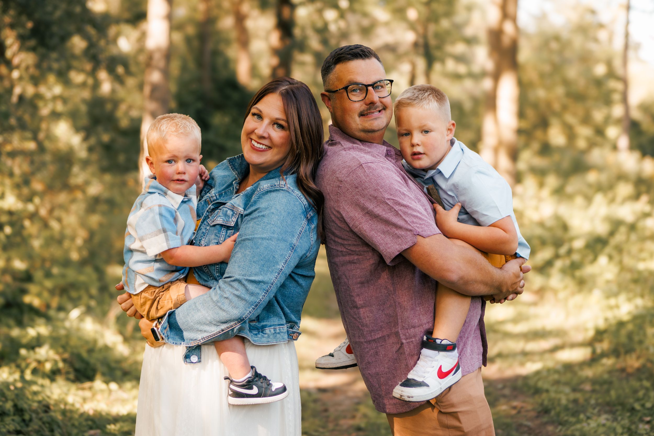 Happy family of four posing outdoors in a sunlit forest, parents holding two young toddler boys, smiling together.
