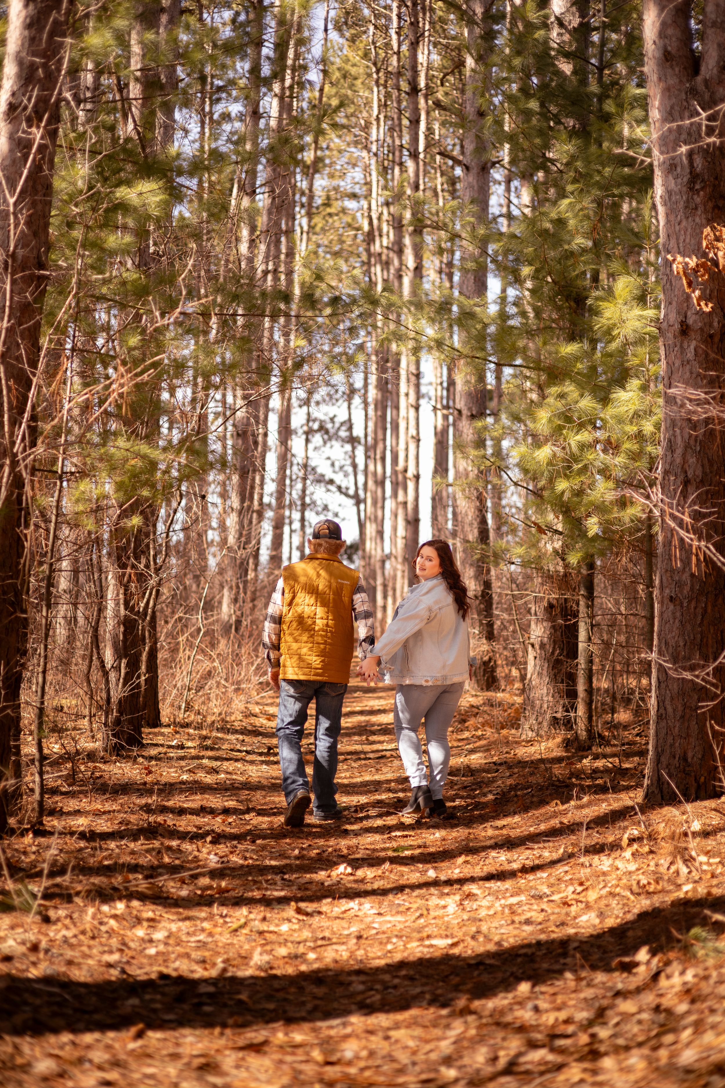 Couple walking hand in hand on a leaf-covered forest path surrounded by tall pine trees in autumn sunlight.