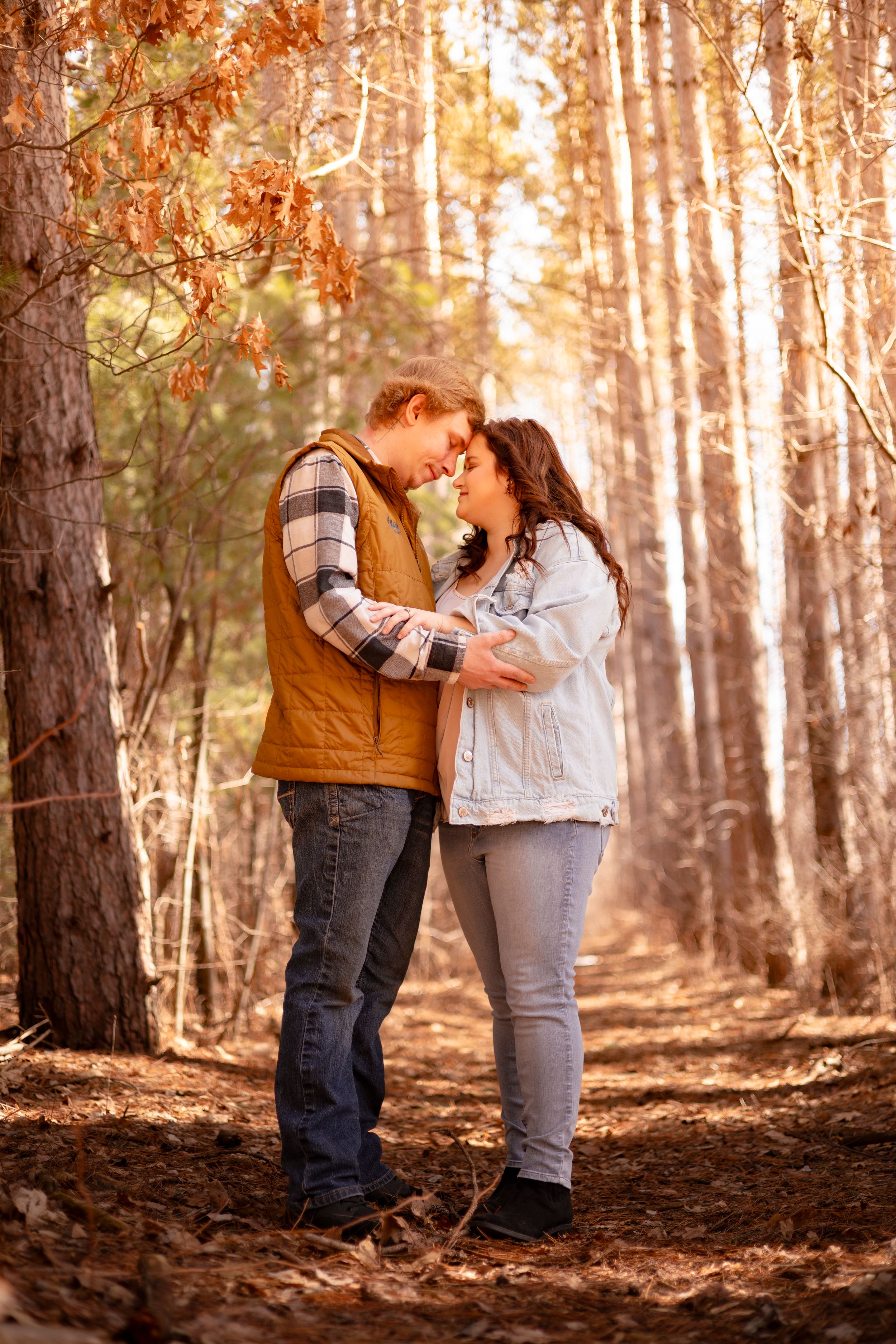 Couple sharing a romantic moment in an autumn forest, surrounded by tall pine trees and golden fallen leaves.