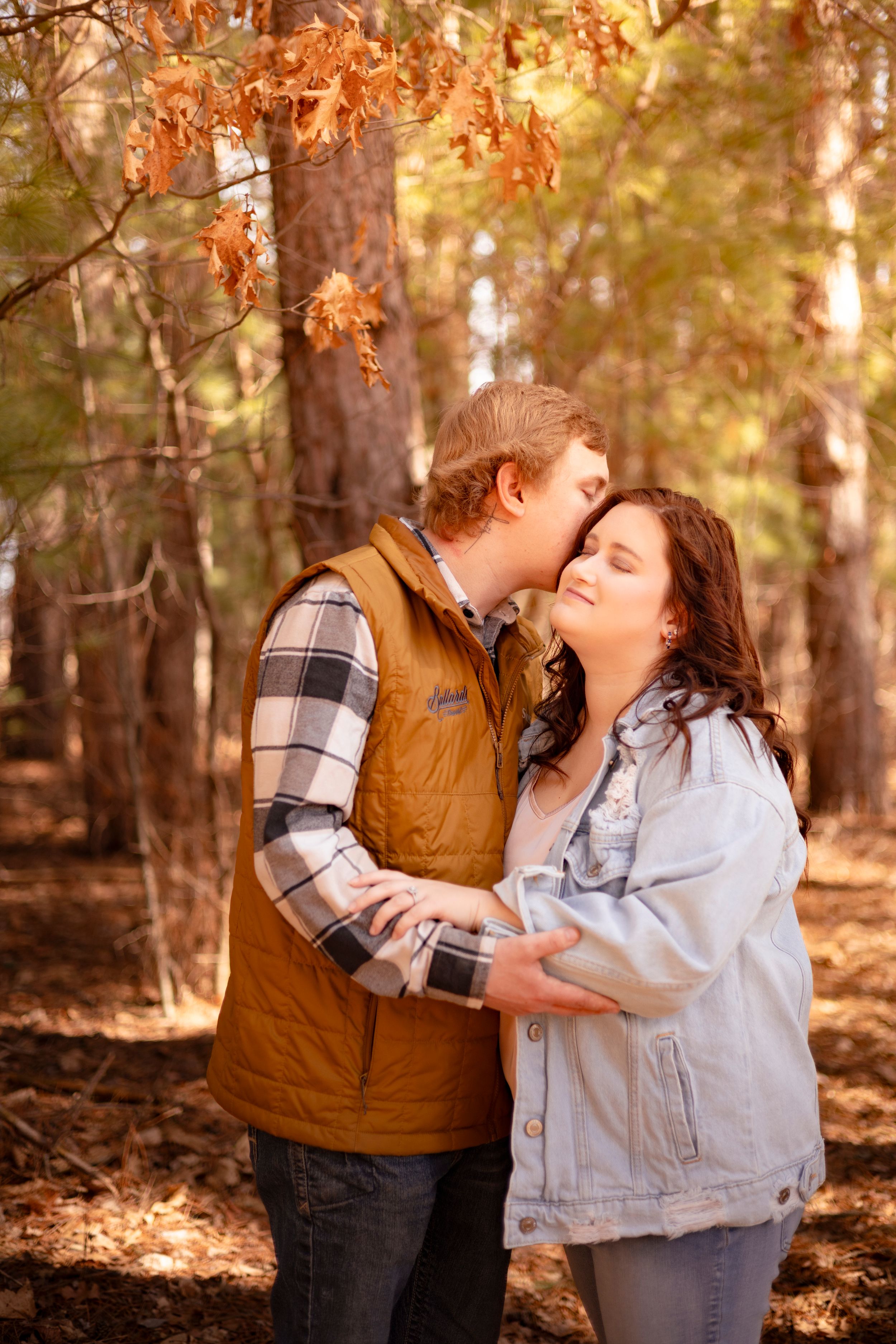 Couple sharing a tender kiss in an autumn forest, woman in denim jacket smiling as man in plaid shirt embraces her.
