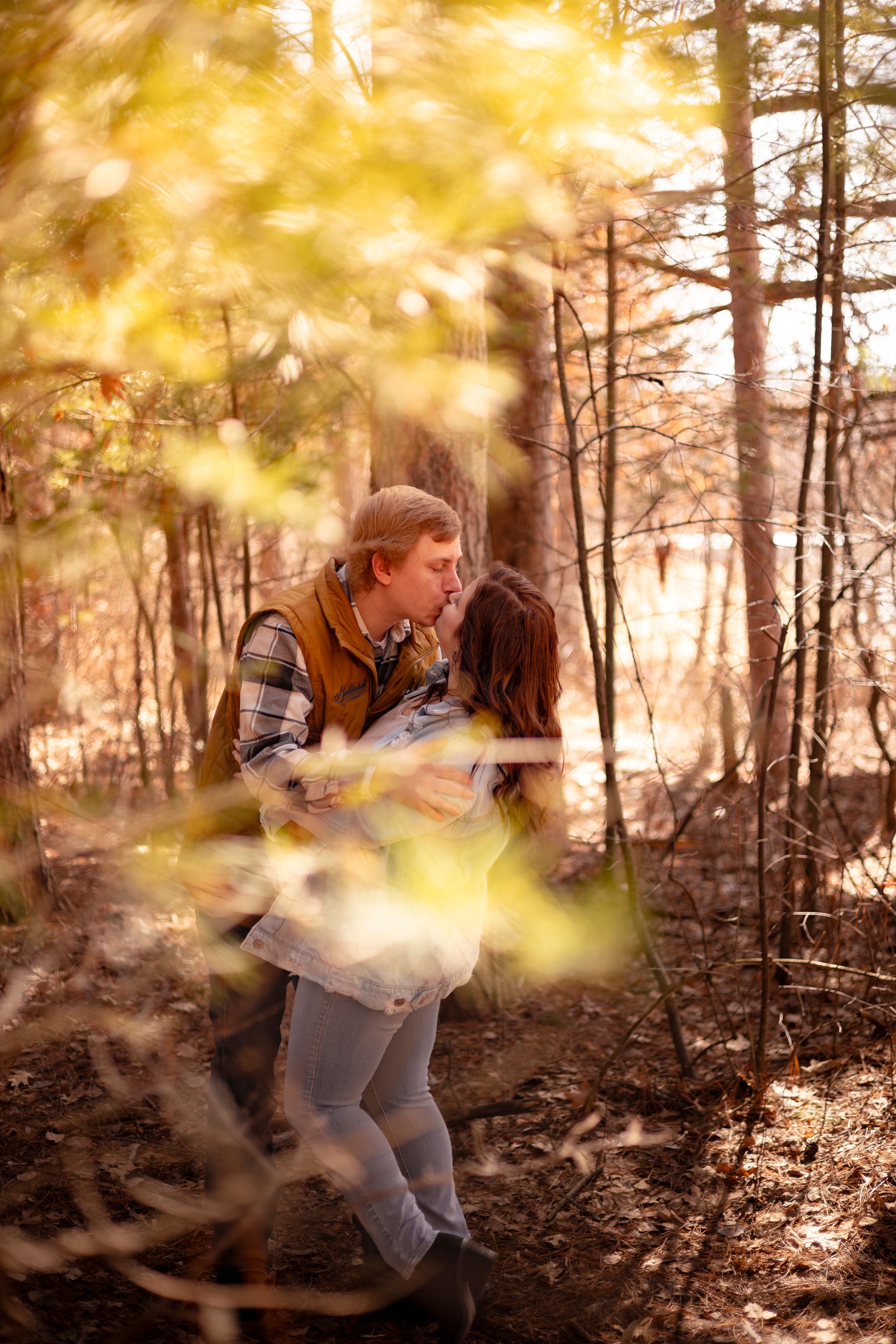 Couple sharing a romantic kiss in a sunlit autumn forest, surrounded by golden leaves and tall trees.