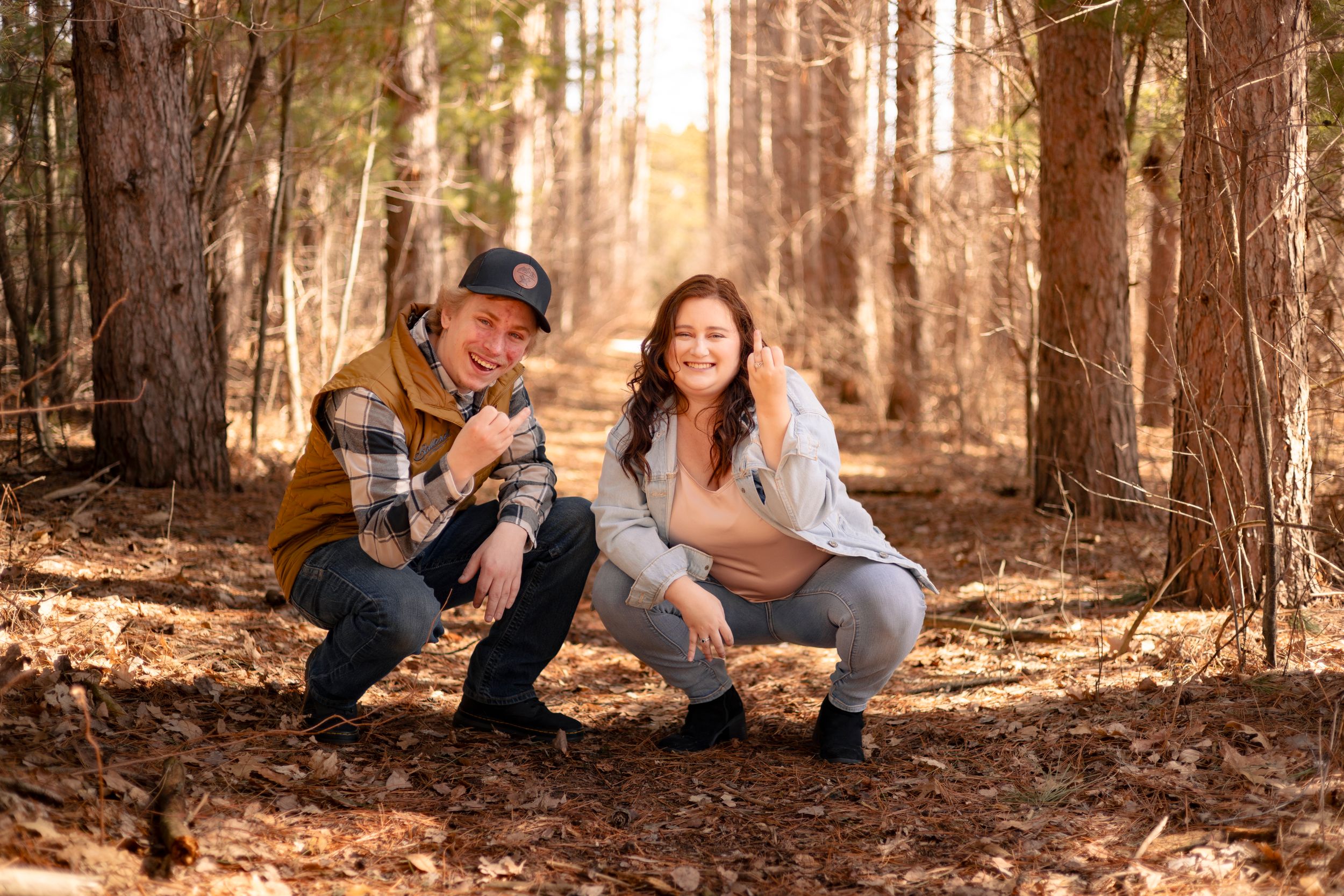 Couple crouching together in a sunlit forest, smiling warmly, woman showing a baby bump among tall pine trees.
