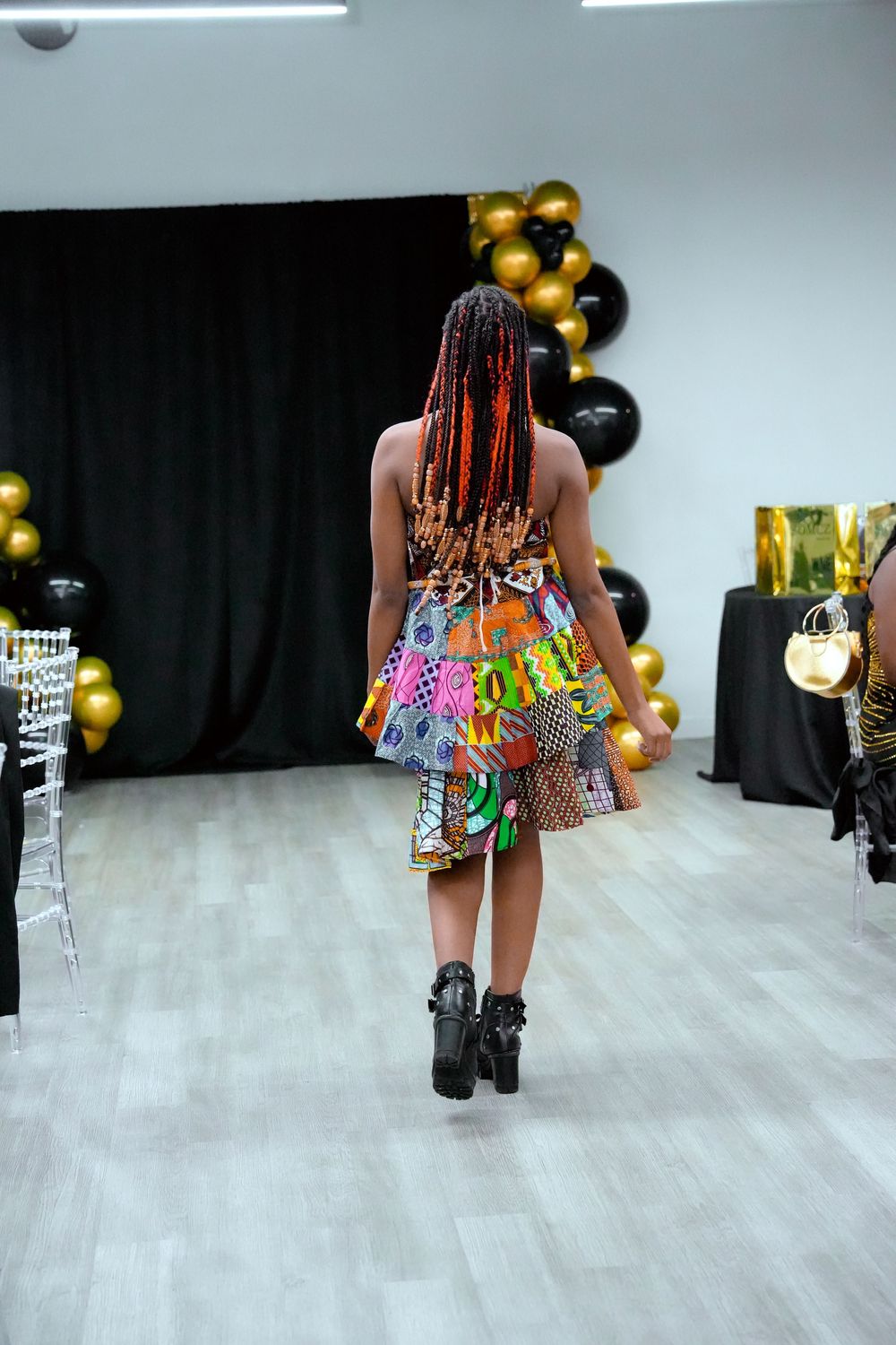 Model walks runway in colorful African print mini dress with red braids, black boots, gold and black balloon decor.