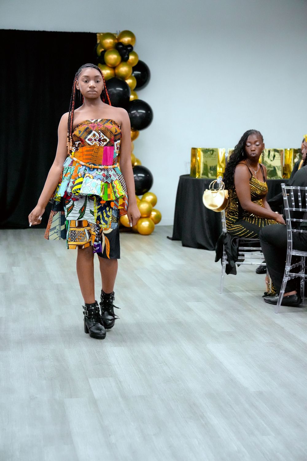 Young model walks runway in colorful African print strapless dress with black boots at fashion show.