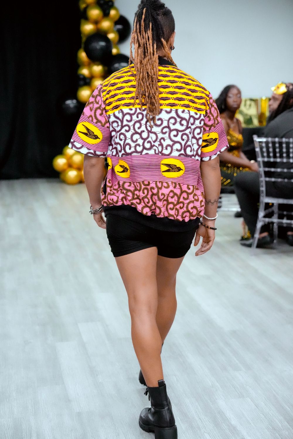 Model walks away on runway wearing vibrant African print patchwork top with black shorts and boots.