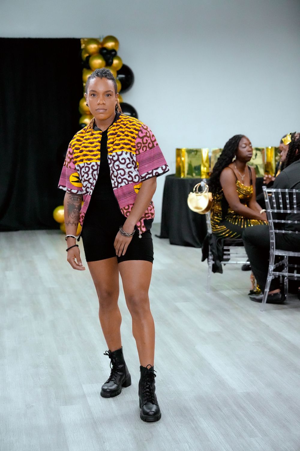 Woman in African print jacket, black shorts, and combat boots poses at a fashion event with gold balloons.