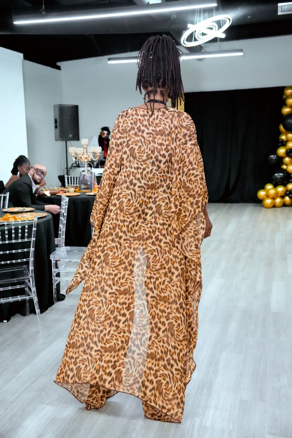 Model walks runway in flowing leopard print maxi dress at fashion show event with gold balloon decor.