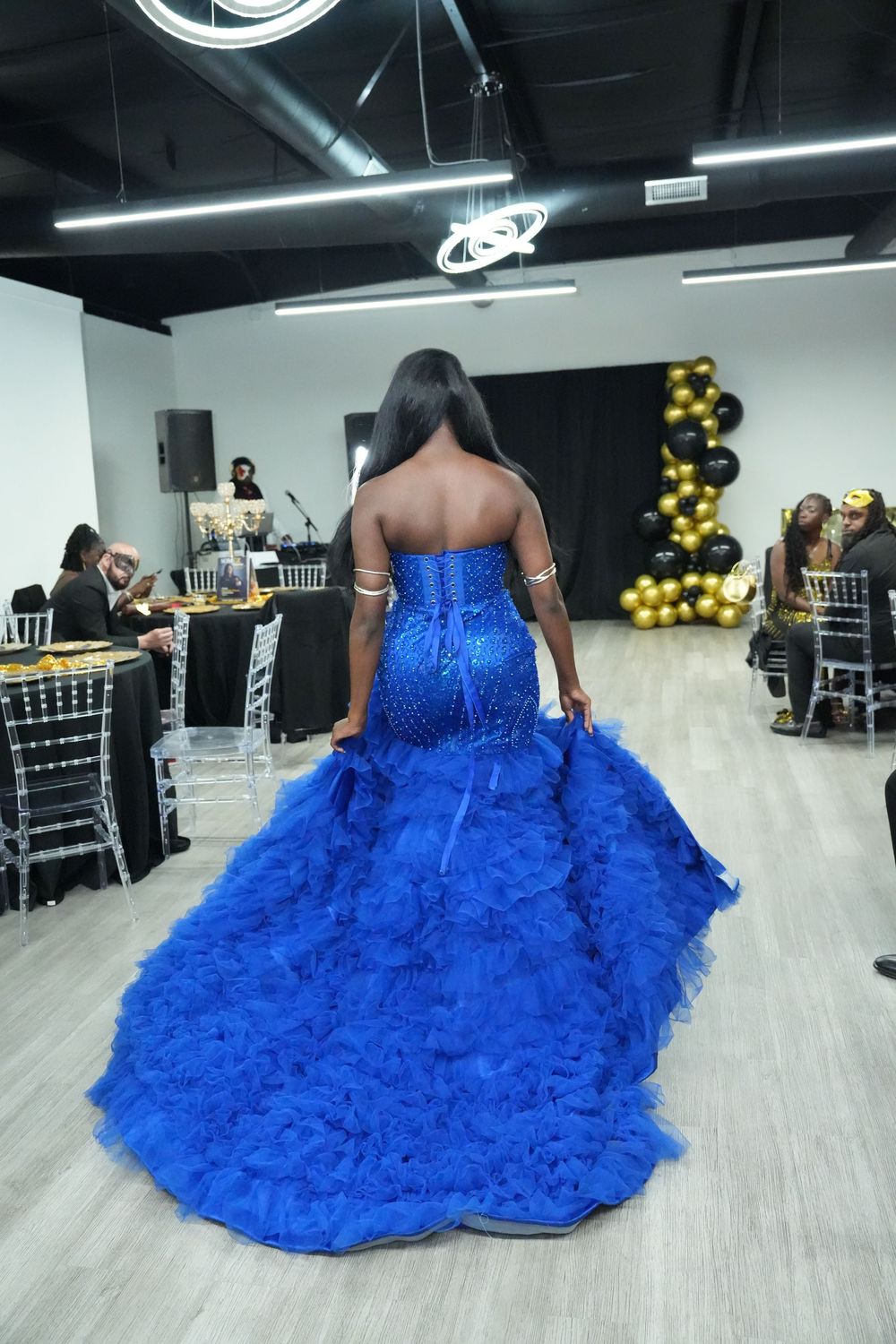 Woman in stunning royal blue ruffled mermaid gown walks runway at elegant event with gold and black decor.