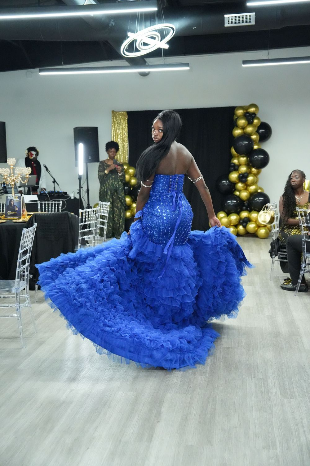 Model walks runway in stunning royal blue ruffled mermaid gown at indoor fashion show with gold and black balloon decor.