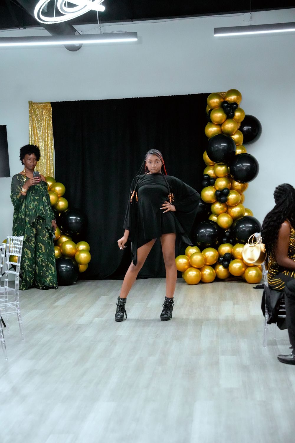 Model poses in black asymmetric mini dress with cape sleeves at fashion show with gold and black balloon backdrop.