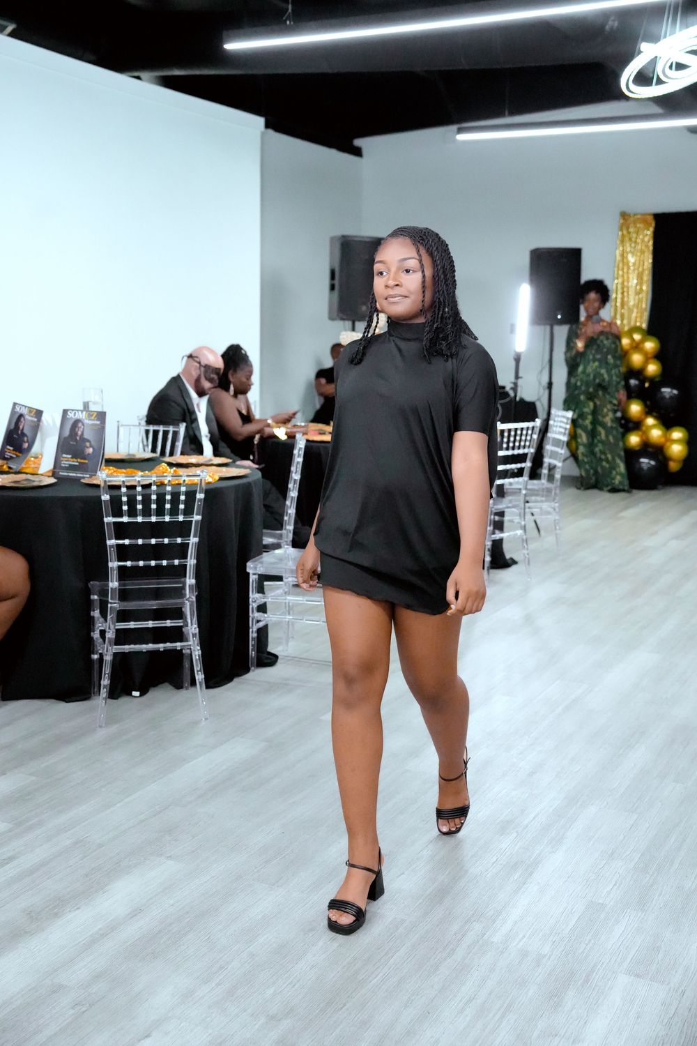 Young woman in black mini dress walks runway at fashion show event with guests seated at decorated tables.