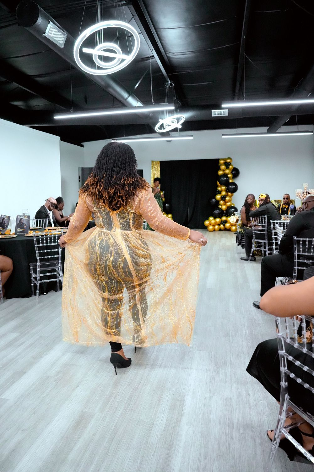 Woman in gold sheer gown walks through elegant event venue with black and gold balloon decor.