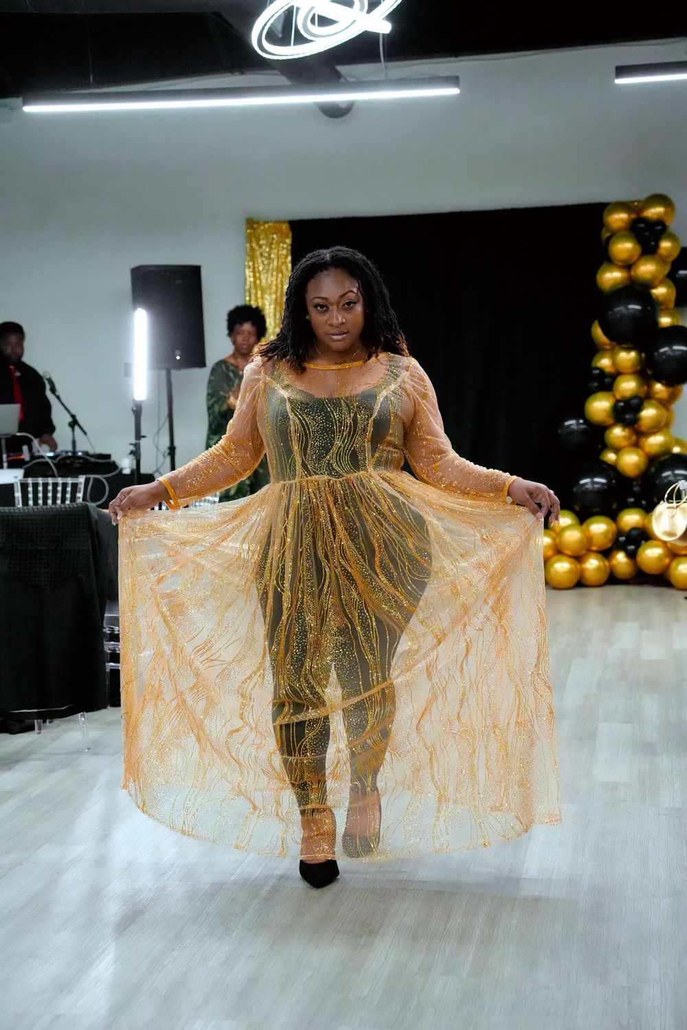 Woman in gold sheer overlay dress posing at fashion show with black and gold balloon decor.