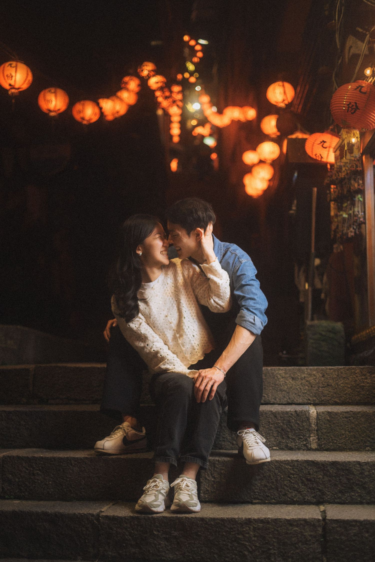 A smiling couple in white outfits sitting on vintage shop steps surrounded by plants and old utility meters.