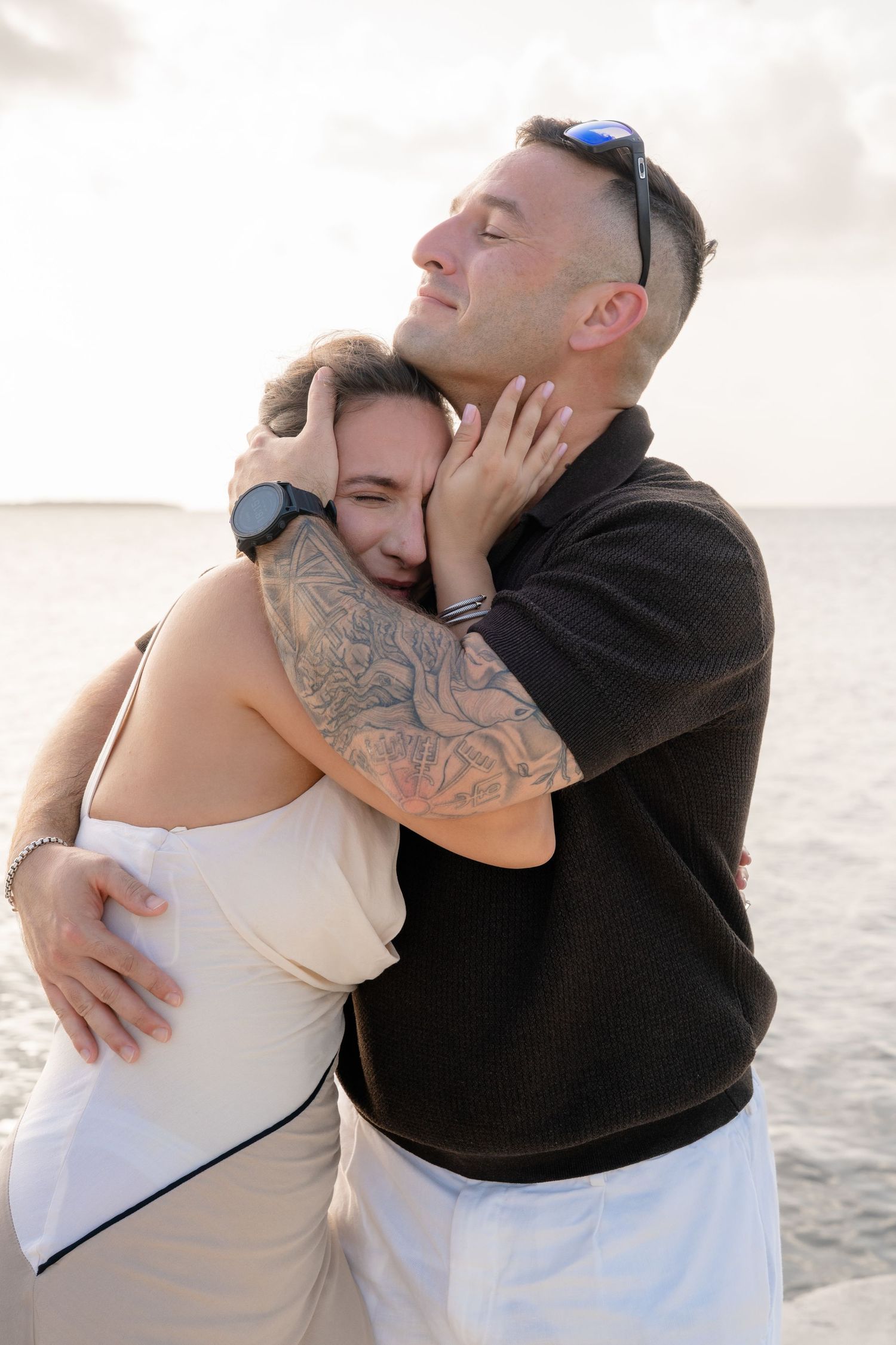 A couple embraces tenderly by the ocean, man with tattoos and sunglasses hugging woman in white dress at sunset.