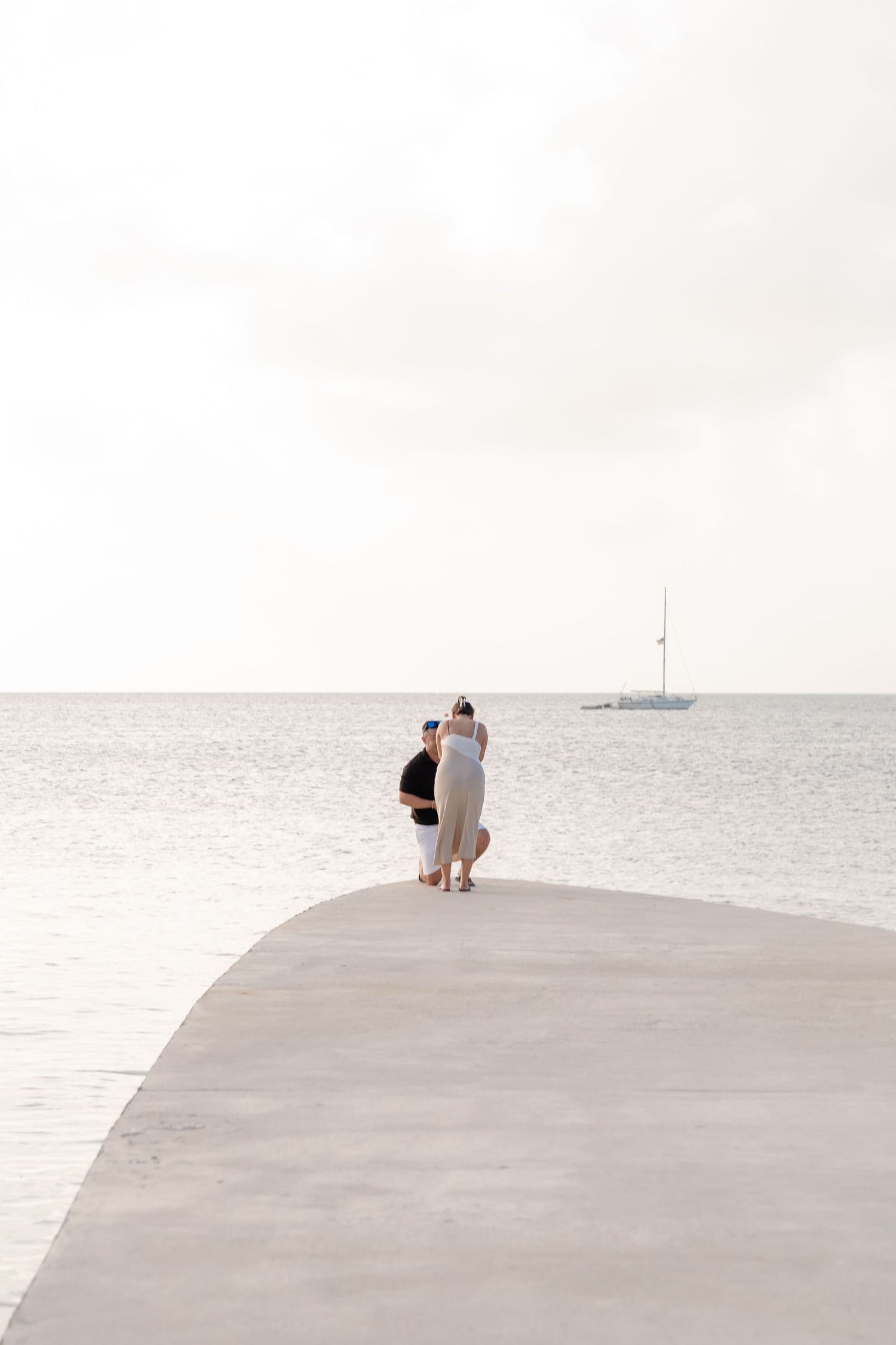 Couple standing on a curved concrete pier overlooking calm ocean waters with a sailboat in the distance.