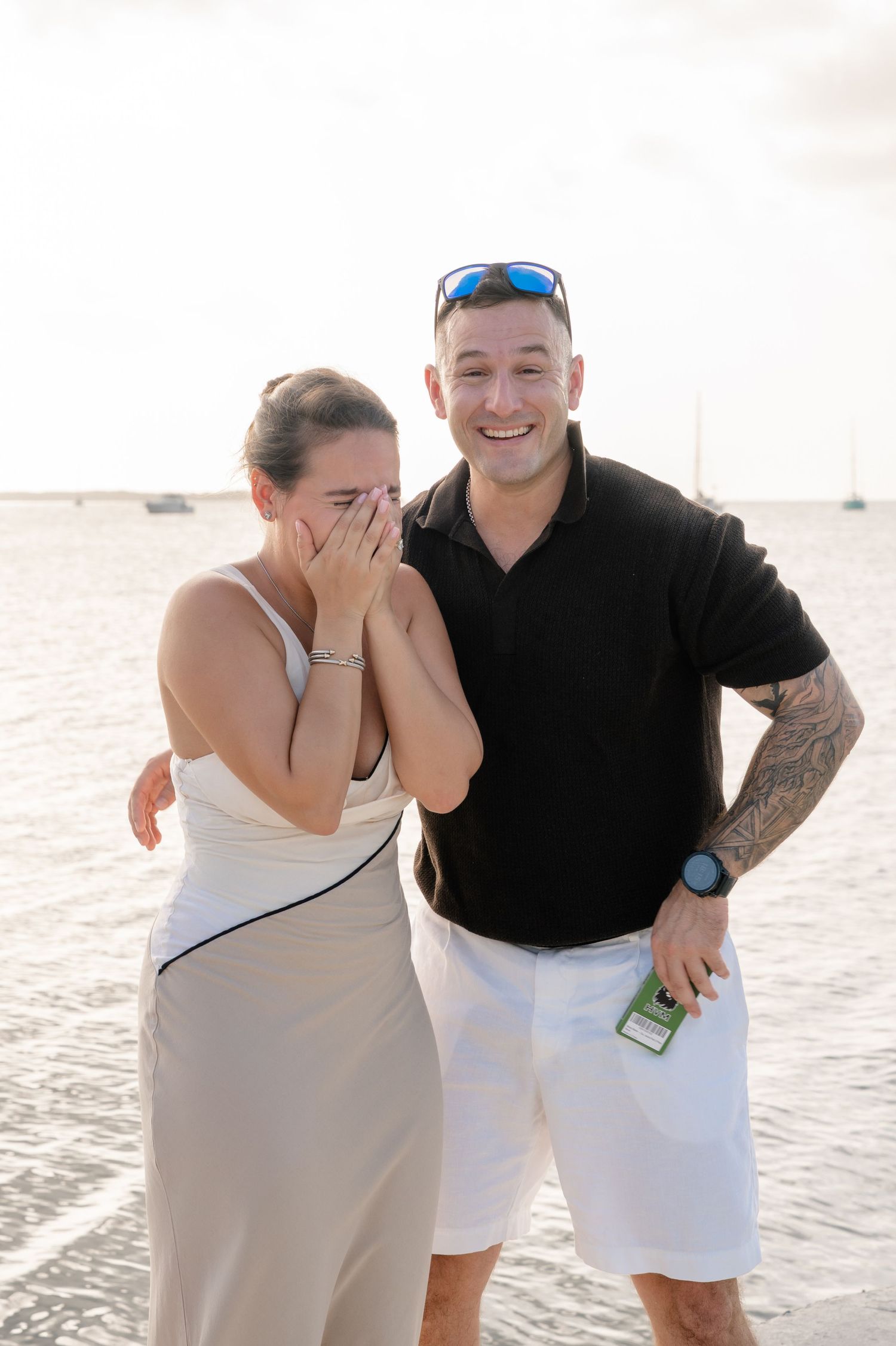 A laughing couple stands on a beach at sunset, woman covering her mouth, man in black polo with tattoos and white shorts.
