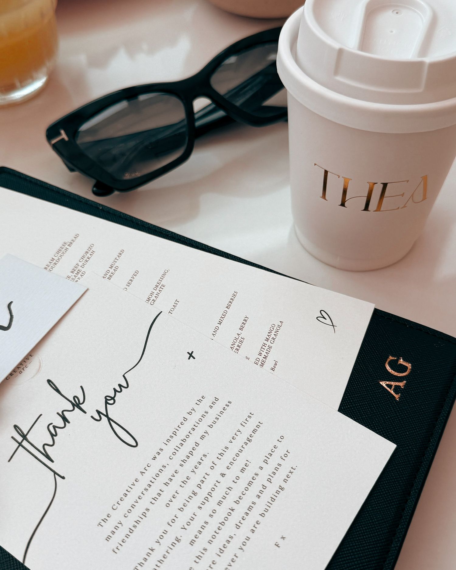 Elegant flat lay with thank you note, AG monogram folder, THEA coffee cup, and black sunglasses on white surface.