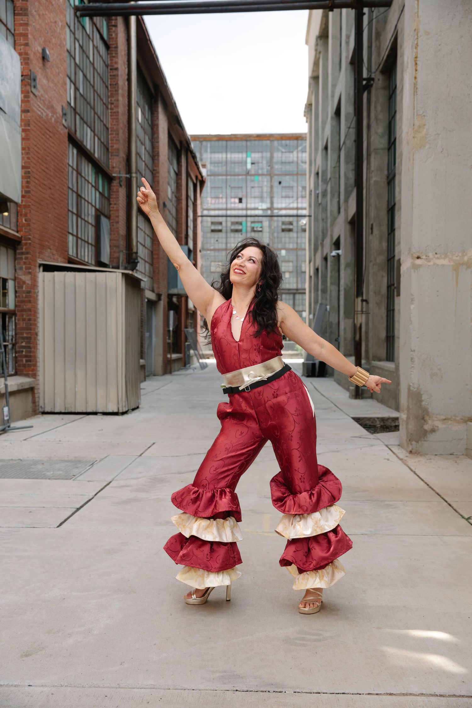 Woman in red ruffled jumpsuit dancing outdoors between urban brick buildings with arms outstretched.