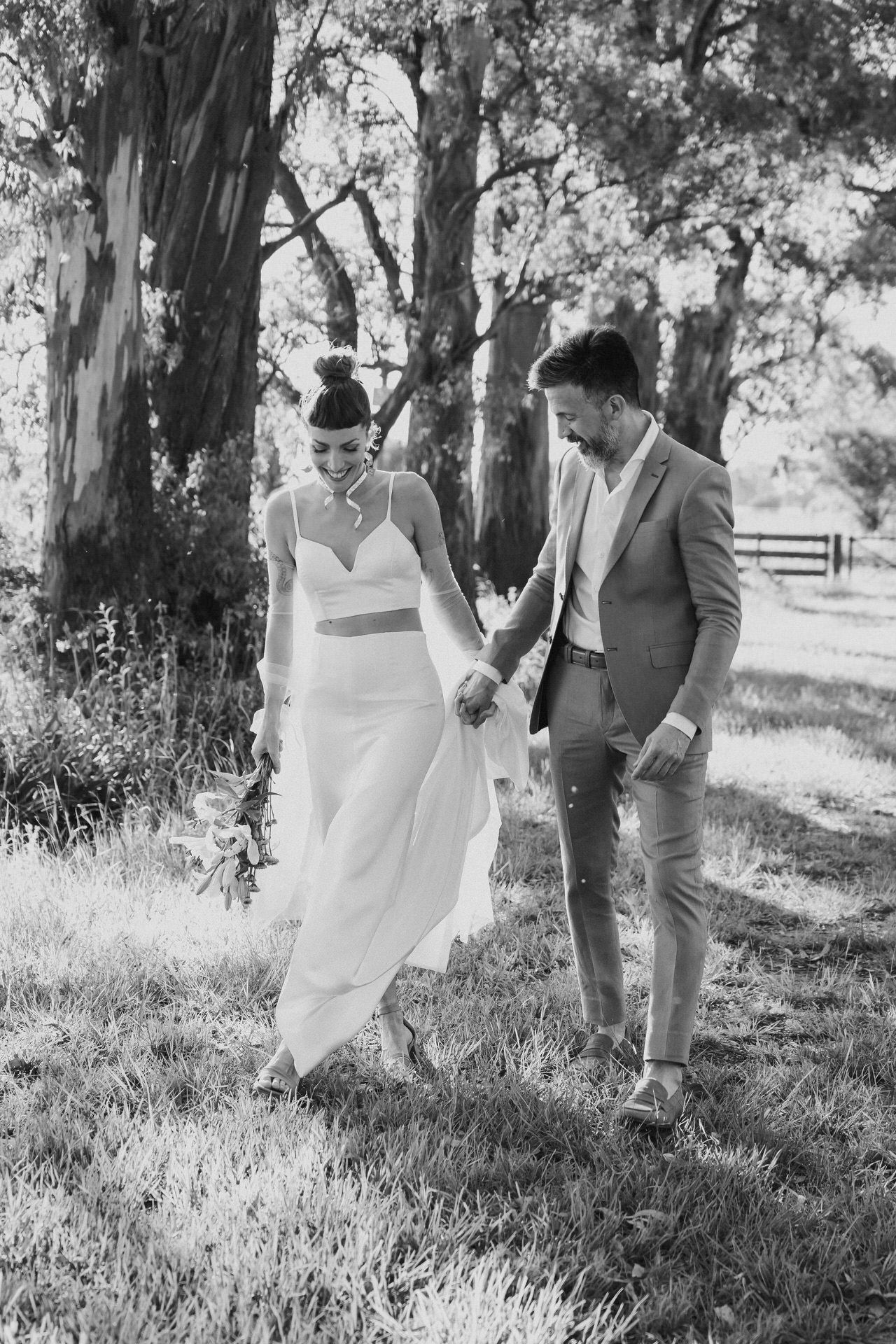 Black and white image of bride and groom walking hand in hand through a scenic tree-lined grassy field.