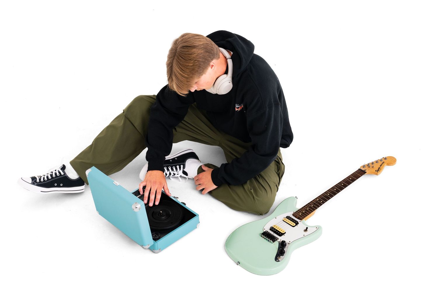 A male model wearing a black hoodie and dark grey jeans sits on the ground touching a blue vinyl player with a light green guitar beside him