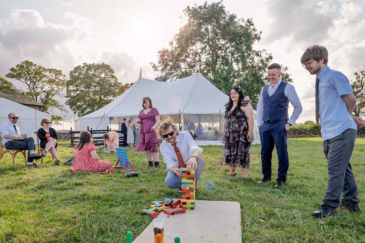 Guests playing Jenga on a lawn outside a white marquee tent at an outdoor wedding reception.