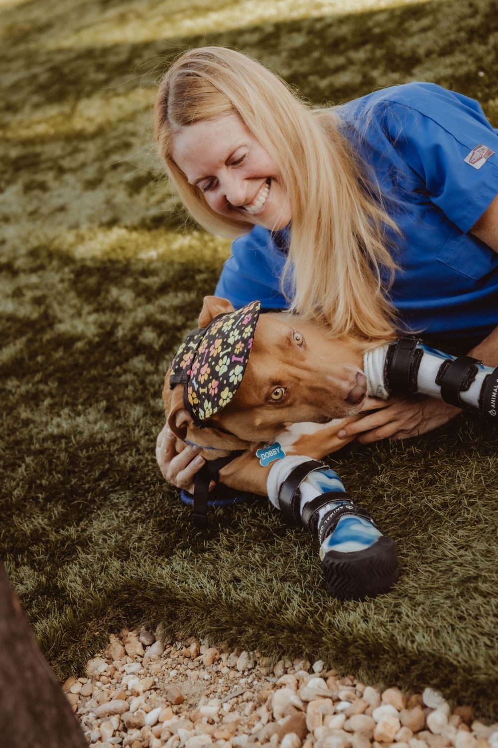 Smiling blonde woman in blue scrubs sitting on grass with a dog wearing colorful boots and a floral bandana.