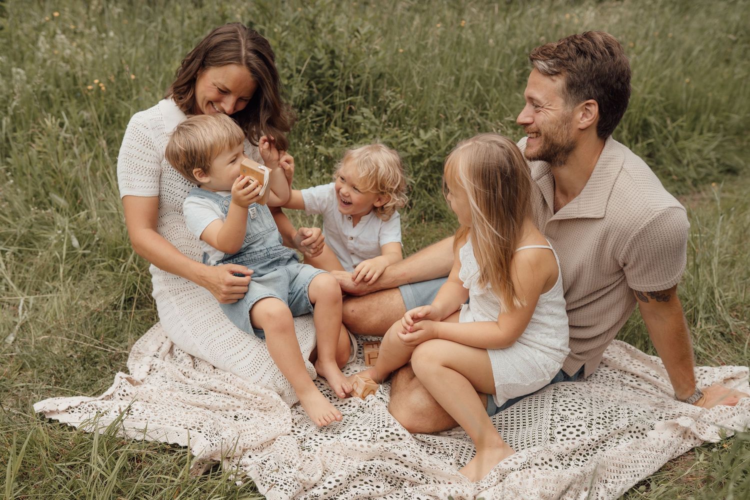 Happy family of five sitting on a blanket in a green meadow, parents smiling with three young children.