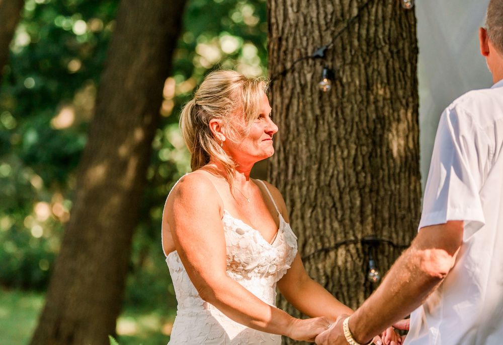 Emotional bride in lace wedding dress holding hands outdoors under trees during wedding ceremony.