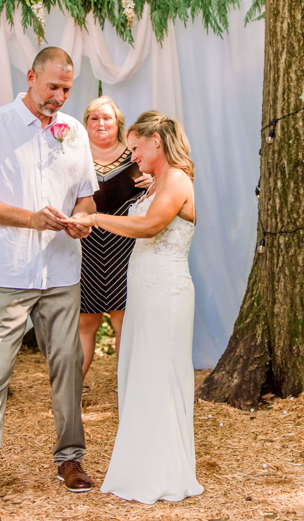 Bride and groom exchange rings during outdoor wedding ceremony with officiant standing behind them near a tree.