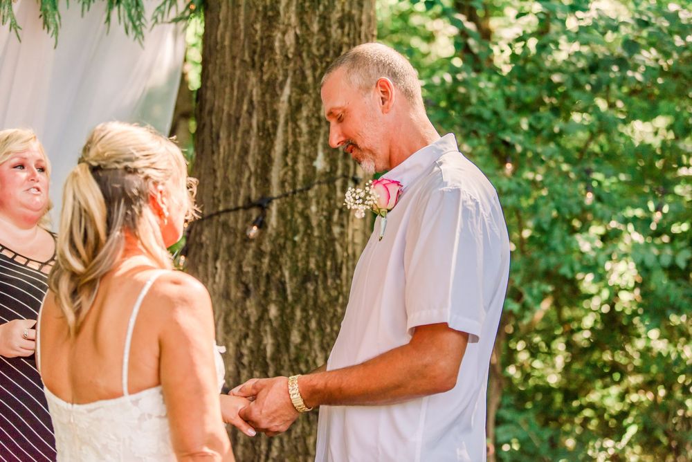 Outdoor wedding ceremony with groom holding bride's hands, surrounded by lush green trees and a floral boutonniere.