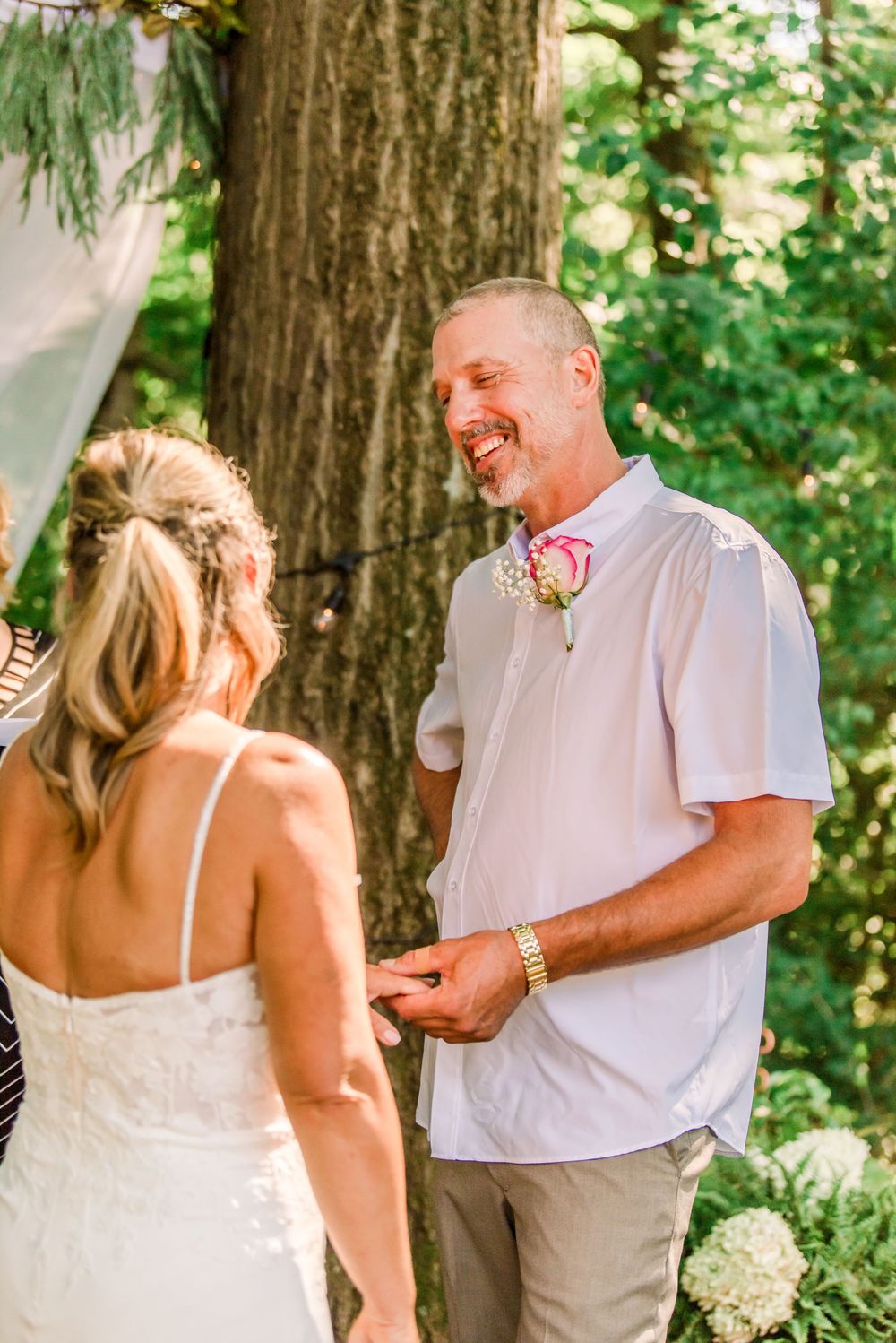 Groom smiling joyfully during outdoor wedding ceremony, wearing white shirt with pink boutonniere, holding bride's hands.
