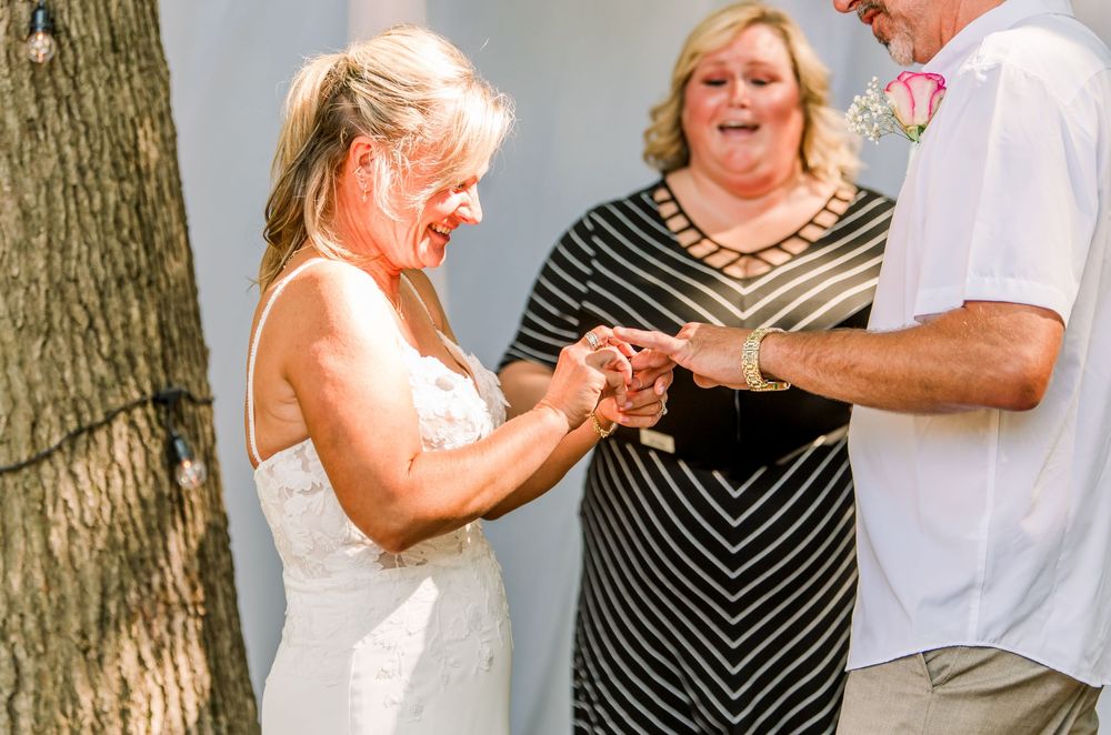 Bride and groom exchanging rings during outdoor wedding ceremony with smiling officiant in black dress.