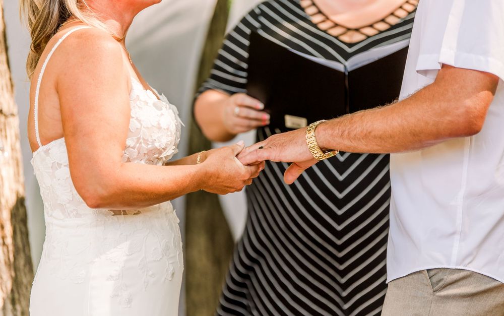 Bride and groom exchanging rings during outdoor wedding ceremony with officiant holding book in background.