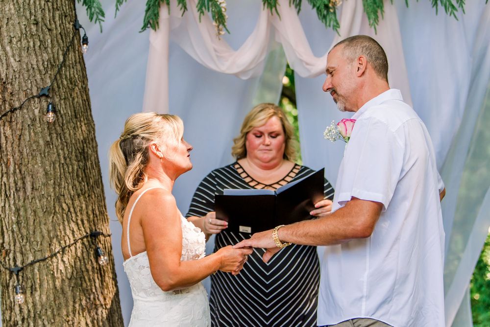 Bride and groom exchange vows outdoors with officiant during intimate wedding ceremony beneath floral arch.