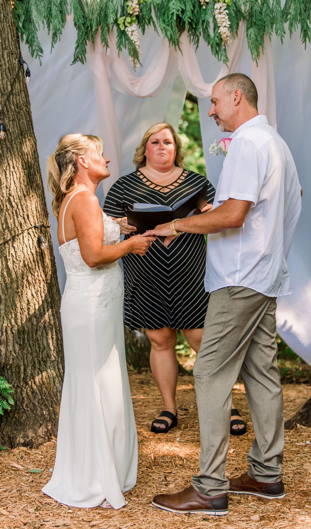 Outdoor wedding ceremony with bride in white gown and groom in white shirt exchanging vows before officiant under floral arch.