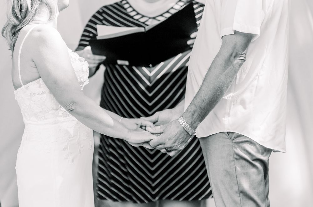 Black and white photo of a couple holding hands during a wedding ceremony with an officiant in a striped dress.
