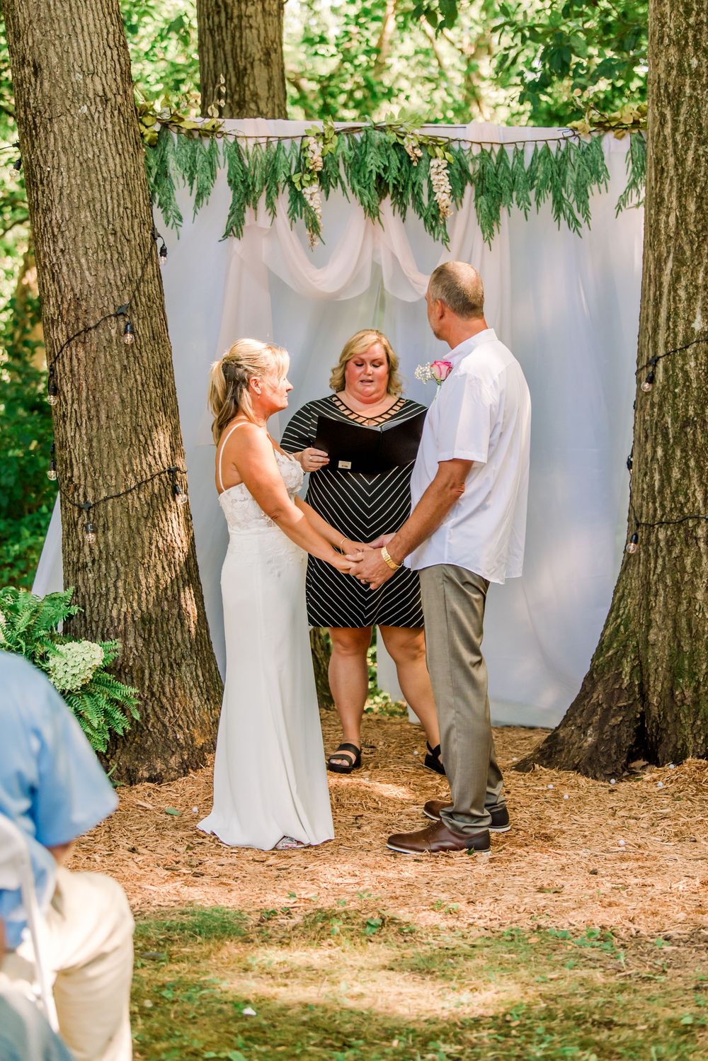 Outdoor wedding ceremony with bride and groom holding hands before officiant, framed by trees and white drape backdrop.