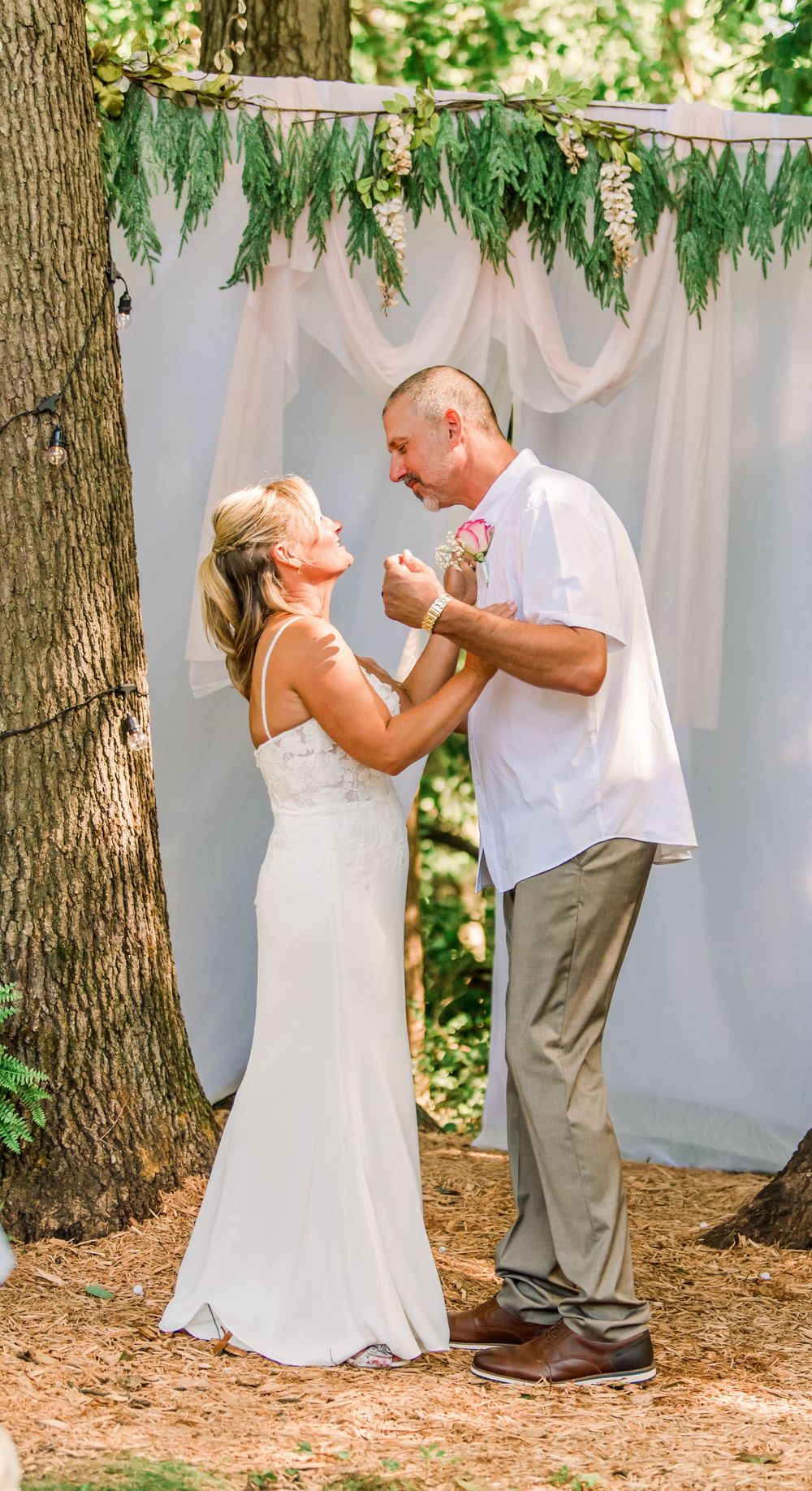 Bride and groom share a joyful first dance outdoors under a rustic floral arch with white draping.