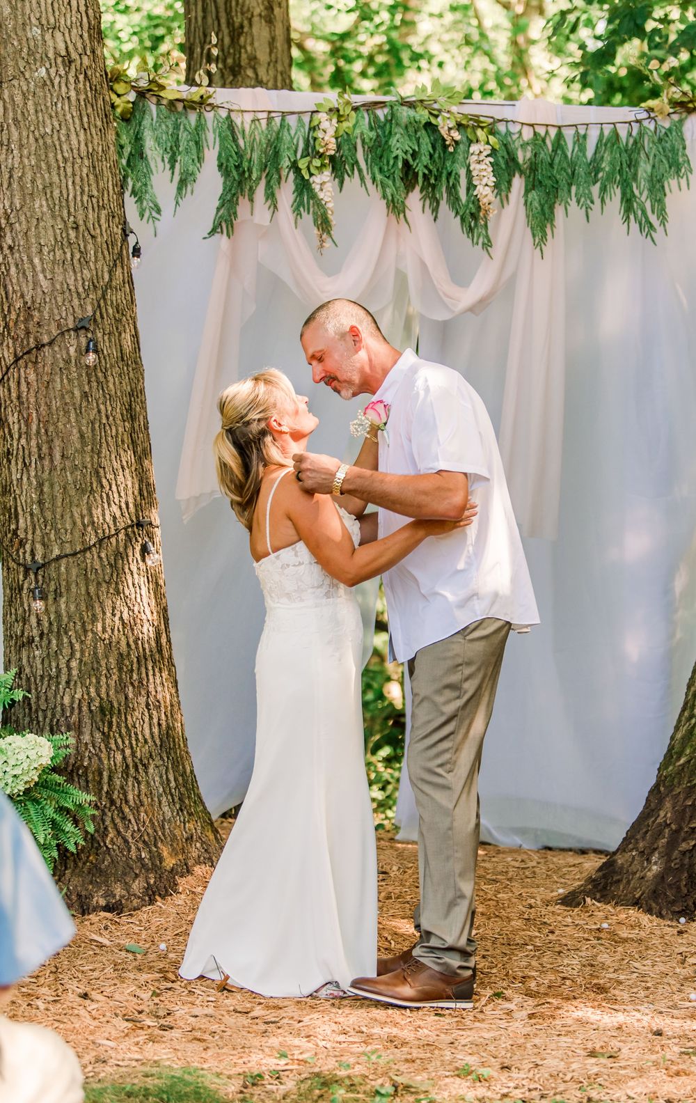 Bride and groom share first dance outdoors under floral arch with white draping at rustic woodland wedding ceremony.