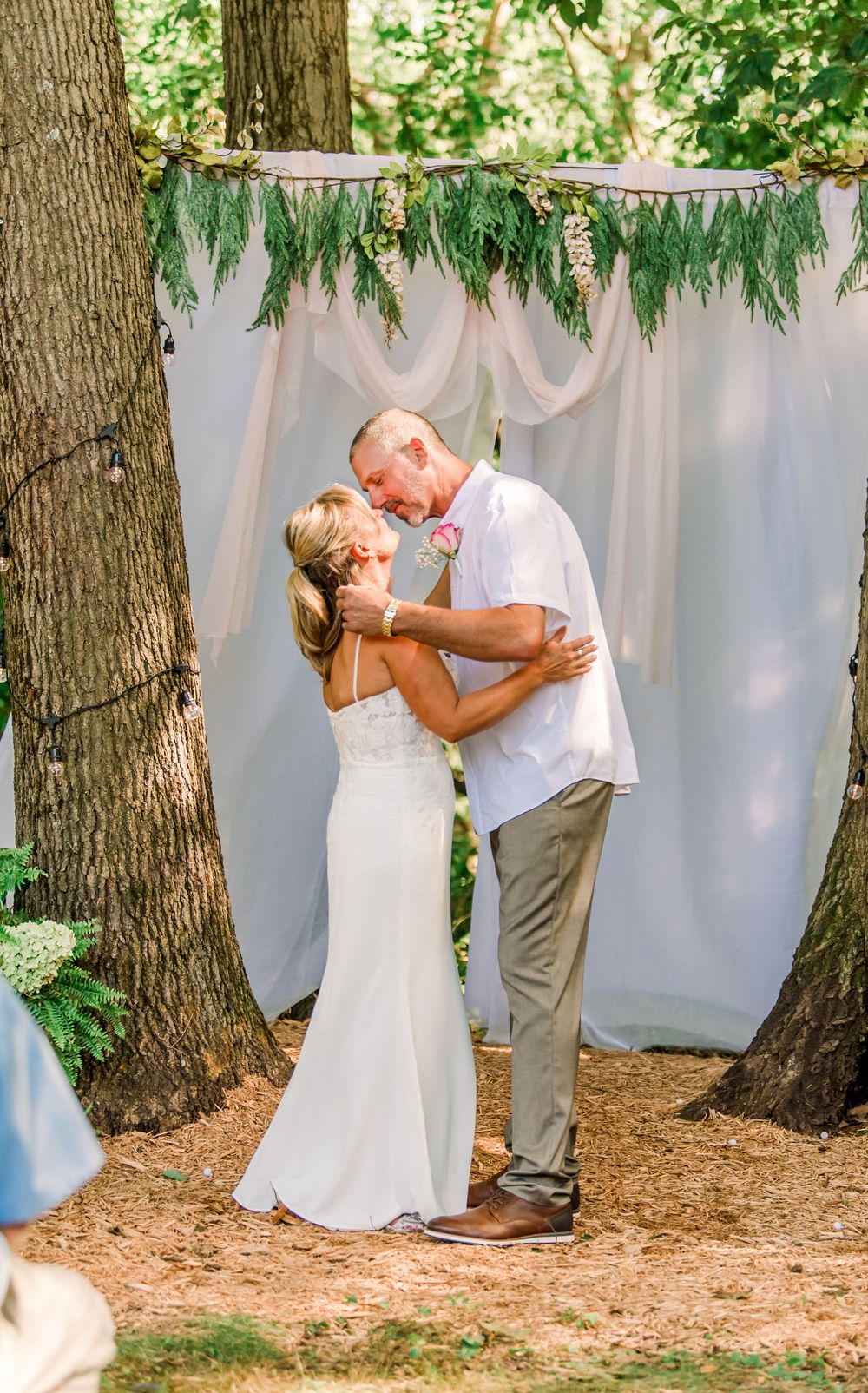 Bride and groom share first kiss at outdoor wedding ceremony beneath rustic greenery arch with white draping.