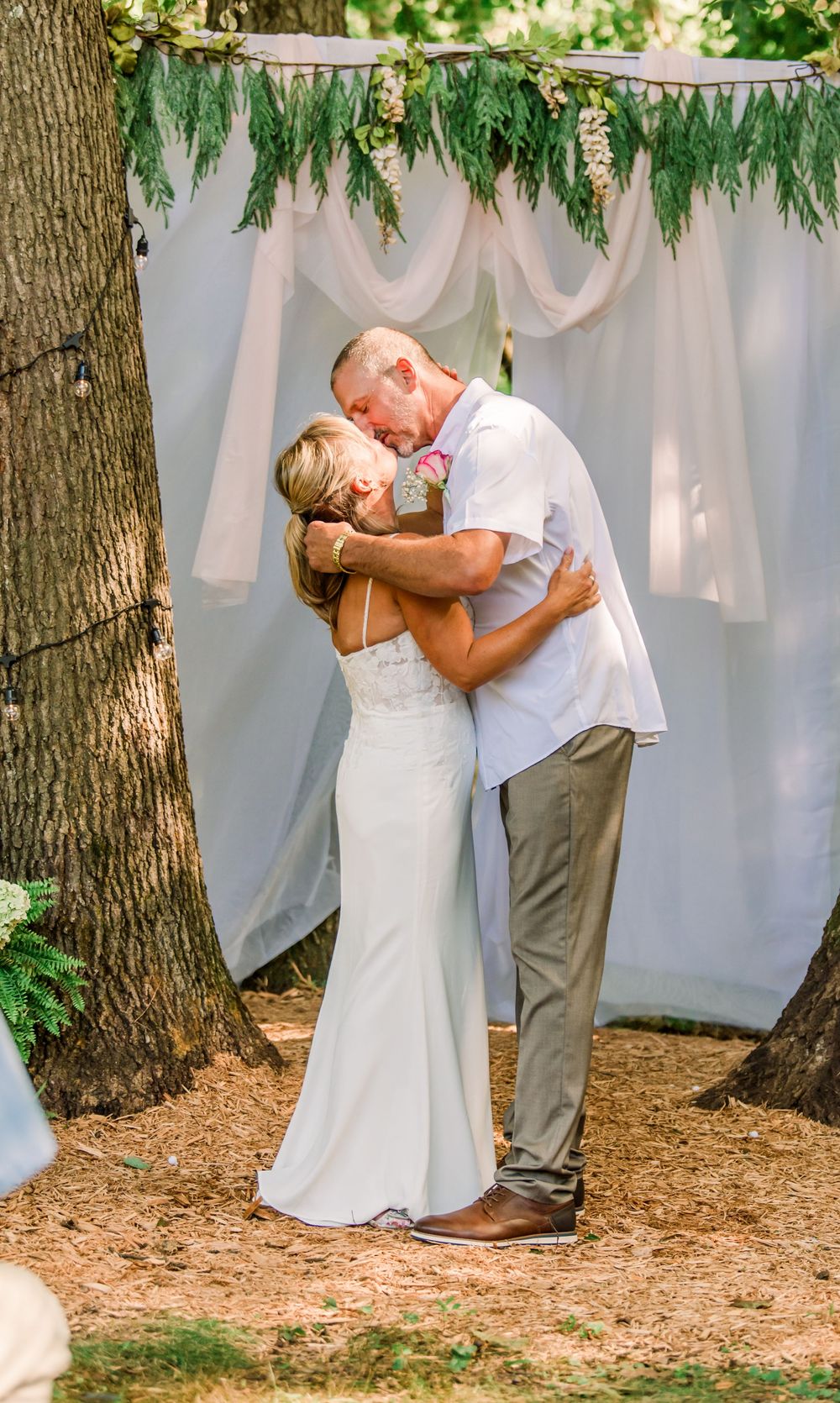 Bride and groom share their first kiss at an outdoor wedding ceremony beneath a white draped arch with greenery.