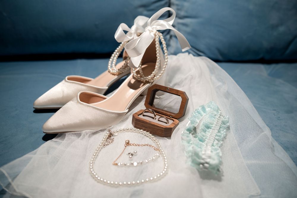 Elegant bridal accessories including white satin heels, pearl necklace, ring box, and lace garter on blue satin.