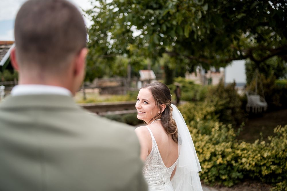 Bride in white gown and veil smiles back at groom in sage suit during outdoor wedding portrait.