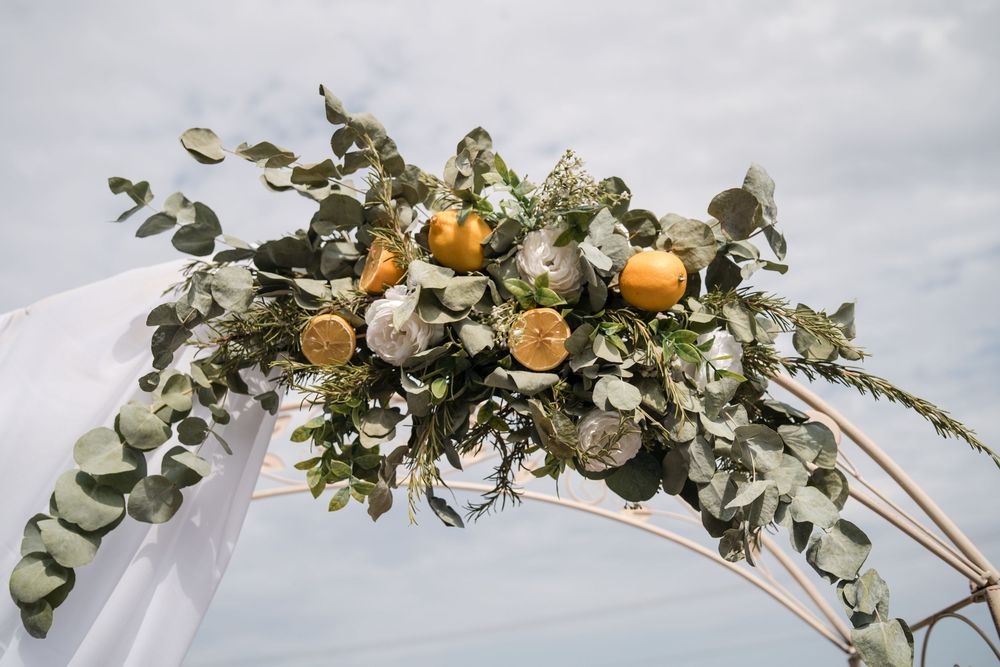 Wedding arch adorned with eucalyptus, white flowers, and golden oranges against a cloudy sky.