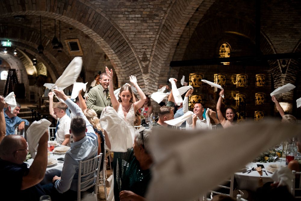 Guests wave white napkins in celebration at a wedding reception in a rustic brick vaulted venue.