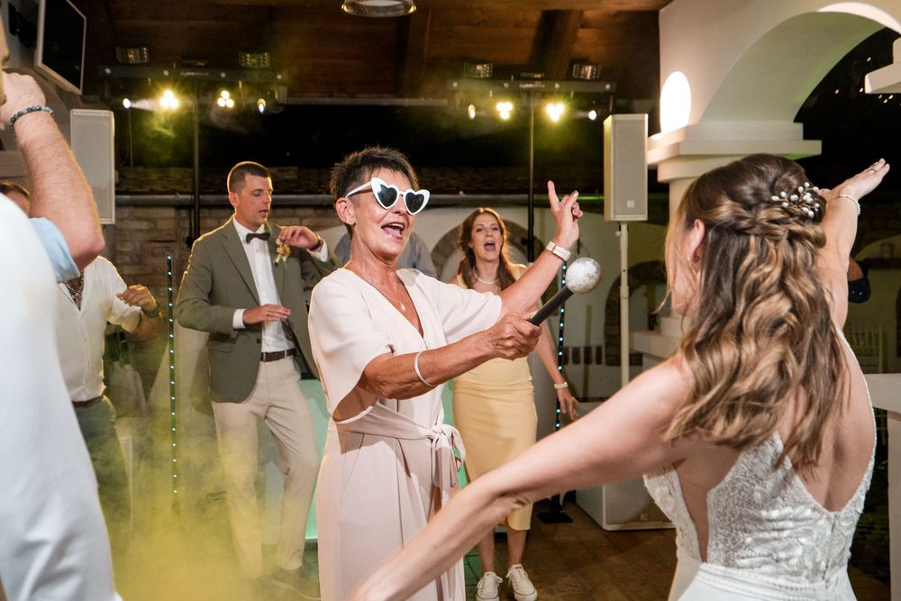 Guests dancing joyfully at a wedding reception, with bride in white gown and friend in funky heart sunglasses.