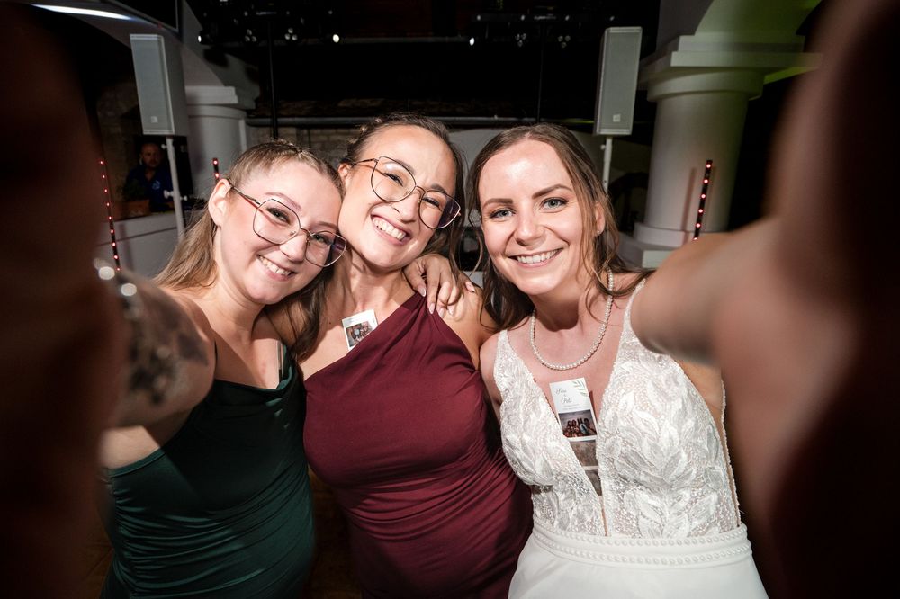 Three smiling women pose for a selfie at a formal event, one wearing a white bridal gown.